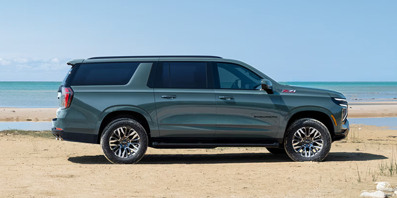 Side profile of a dark green Suburban parked on a sandy beach with the ocean in the background under a clear sky.