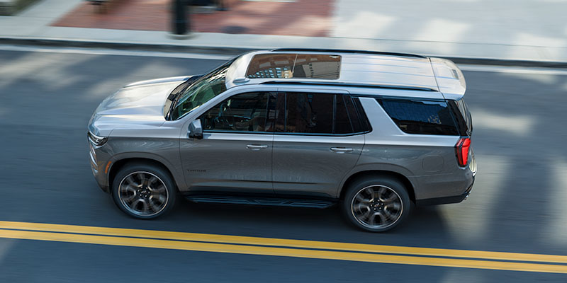 A silver Tahoe driving on a city road captured from an elevated angle, showing motion blur on the street and a modern roof design.